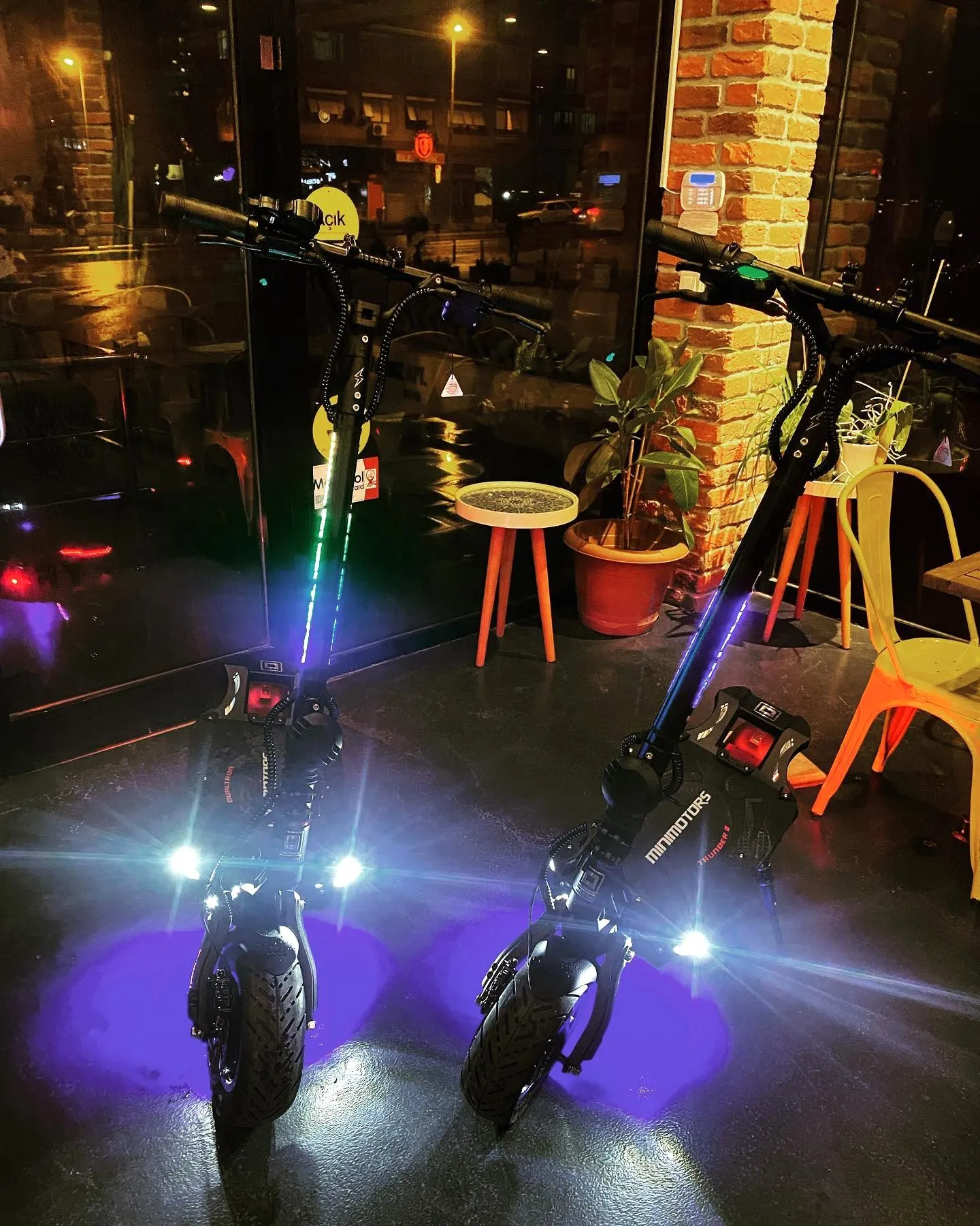 Gece cam cepheli bir mekânda farları ve mor zemin ışıkları yanan iki Dualtron scooter