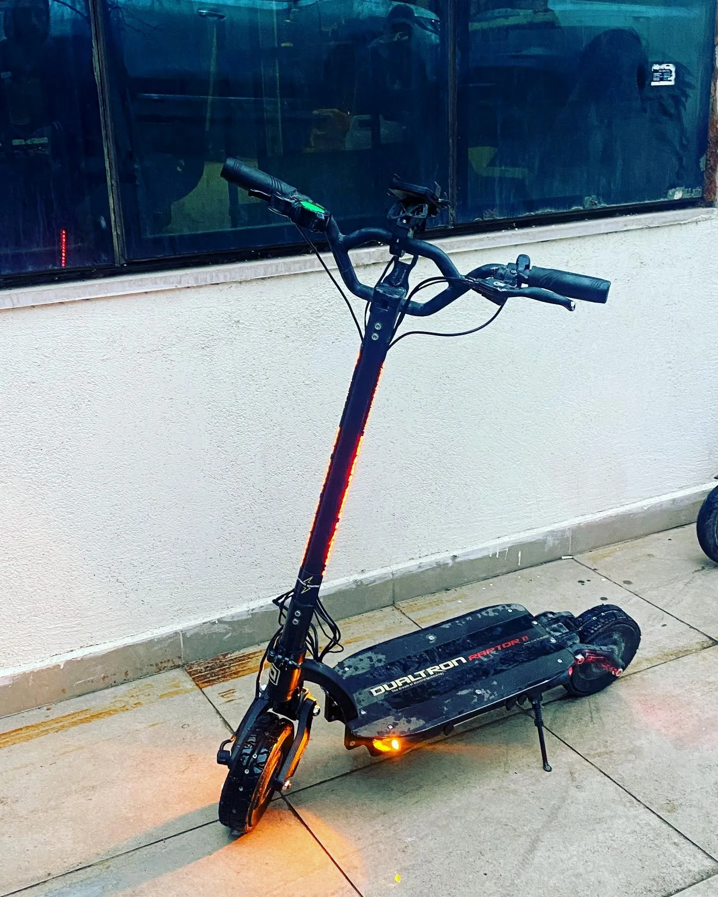 Gövdesinde ve gidonunda turuncu LED yanan Dualtron scooter, beyaz duvar ve cam önünde yan ayağında duruyor