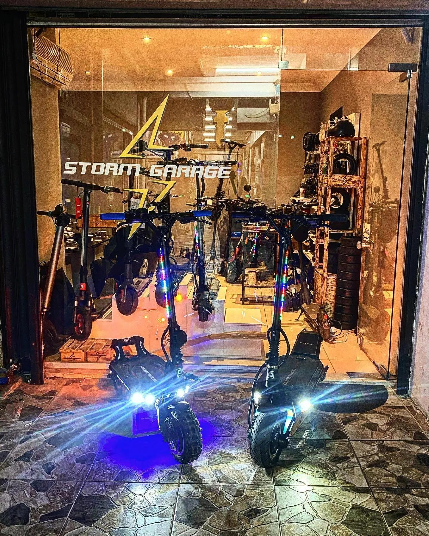 Gece eski Storm Garage dükkânı: camda parlayan şimşekli logo, kapıda farları yanan iki Dualtron, içeride sıralı scooterlar