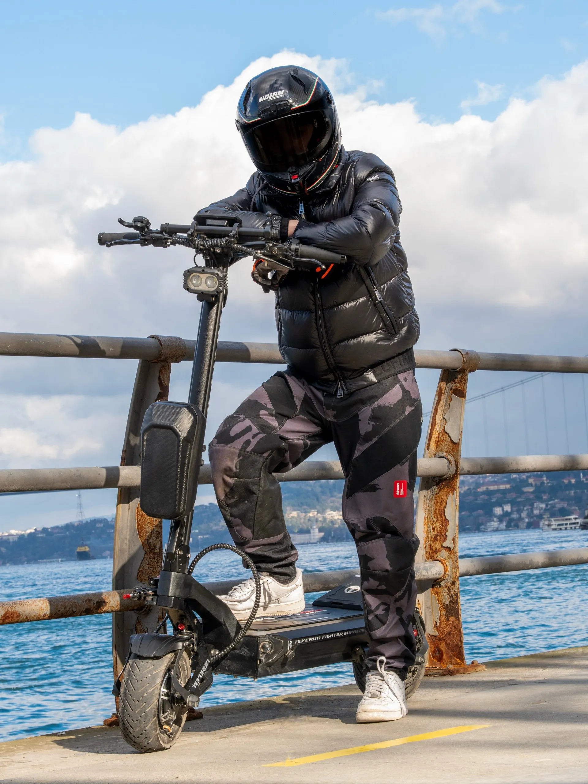 Siyah kask ve şişme montlu sürücü, Boğaz kıyısında paslı korkuluğun önünde Teverun Fighter Supreme scooter'ına yaslanıyor