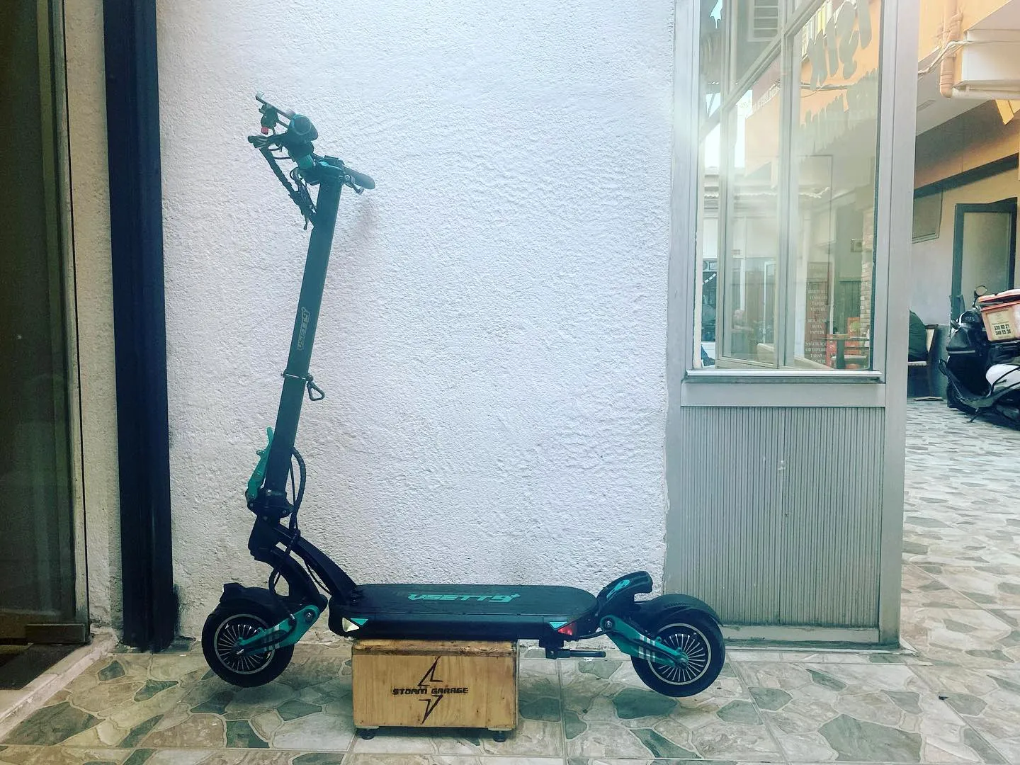 Turkuaz detaylı VSETT 9+ scooter, Storm Garage logolu ahşap kasa üzerinde beyaz duvar önünde yandan